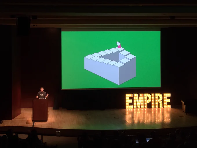 Speaker on stage with slide from his talk with the Penrose stairs on it