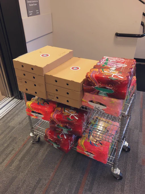A metal cart with wheels that has three shelves. The cart is completely full of boxes of donuts and LaCroix soda. It is in front of an open elevator.