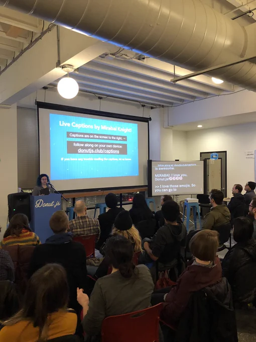 A person giving a talk at a podium in front to a room full of people in chairs. Behind the speaker is a projector screen with instructions for watching the captions on your own screen. The screen for captions is visible as well.