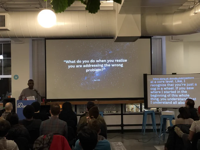 A person giving a talk at a podium in front to a room full of people in chairs. Behind the speaker is a projector screen with a quote: “What do you do when you realize you are addressing the wrong problem?”