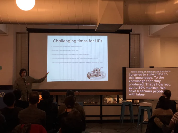 A person giving a talk at a podium in front to a room full of people in chairs. Behind the speaker is a projector screen with the title ‘Challenging times for University Presses.’