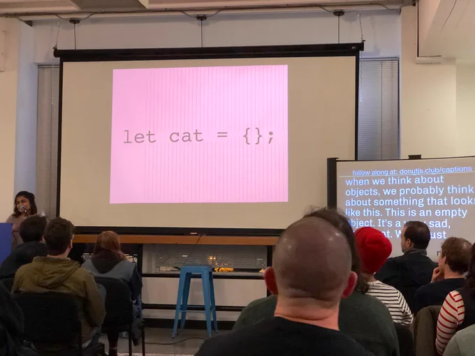 A person giving a talk at a podium in front to a room full of people in chairs. Behind the speaker is a projector screen with code on it: let cat = {};