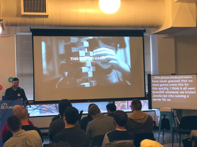 A person giving a talk at a podium in front to a room full of people in chairs. Behind the speaker is a projector screen with the title “This stuff is hard.”