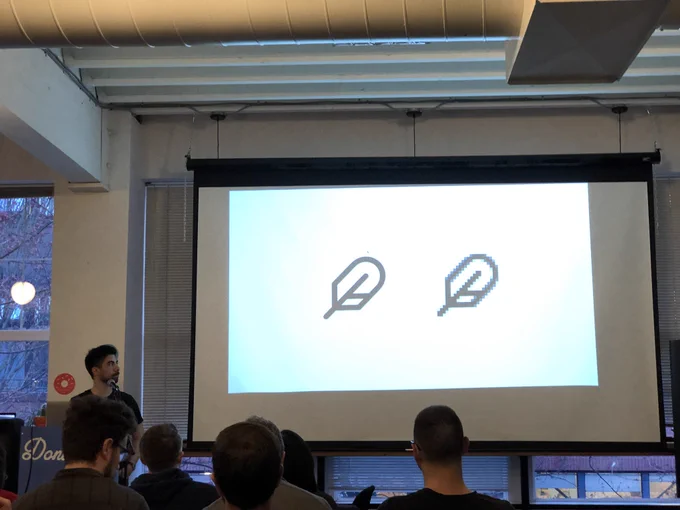 A person giving a talk at a podium in front to a room full of people in chairs. Behind the speaker is a projector screen with two feather icons. One is smooth and the other is pixelated.