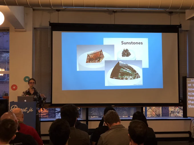 A person giving a talk at a podium in front to a room full of people in chairs. Behind the speaker is a projector screen with photos of gemstones.