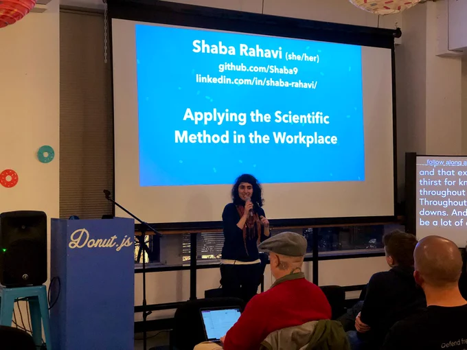A person giving a talk at a podium in front to a room full of people in chairs. Behind the speaker is a projector screen with the speaker’s name and talk title: Shaba Rahavi, Applying the Scientific Method in the Workplace