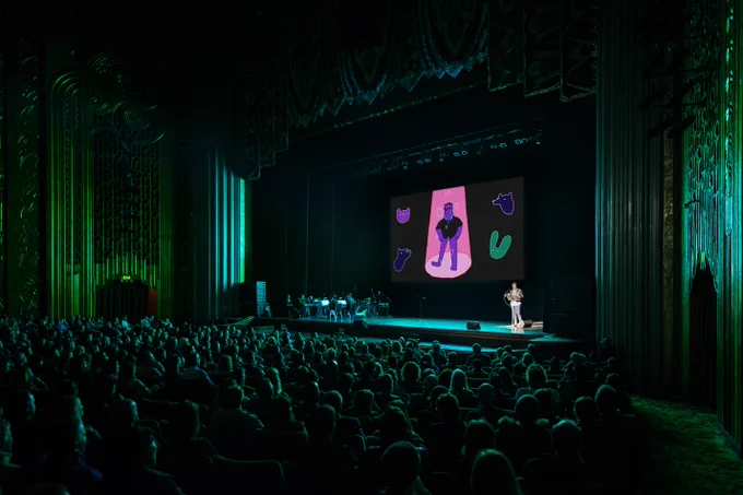 a crowd in a theater watches a person giving a presentation on a stage. there are illustrations behind the presenter on a large screen. there is a band assembled on the opposite side of the stage.
