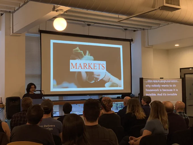 A person giving a talk at a podium in front to a room full of people in chairs. Behind the speaker is a projector screen reading “MARKETS”