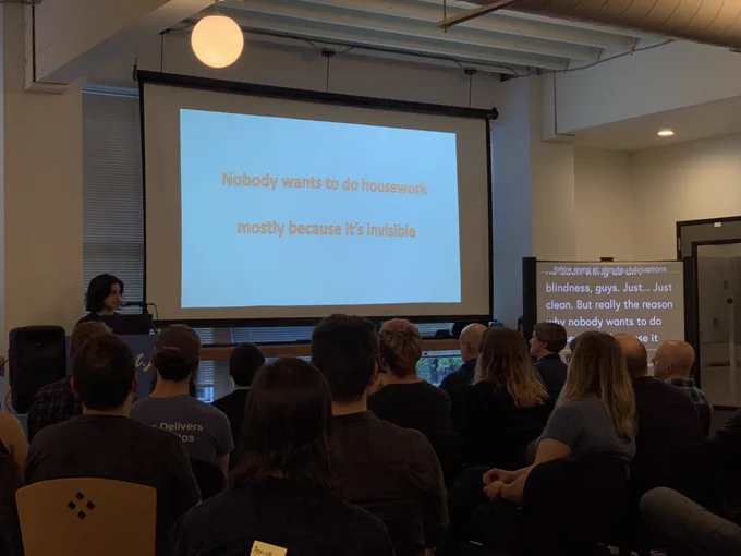 ‪A person giving a talk at a podium in front to a room full of people in chairs. Behind the speaker is a projector screen reading “nobody wants to do housework mostly because it’s invisible”