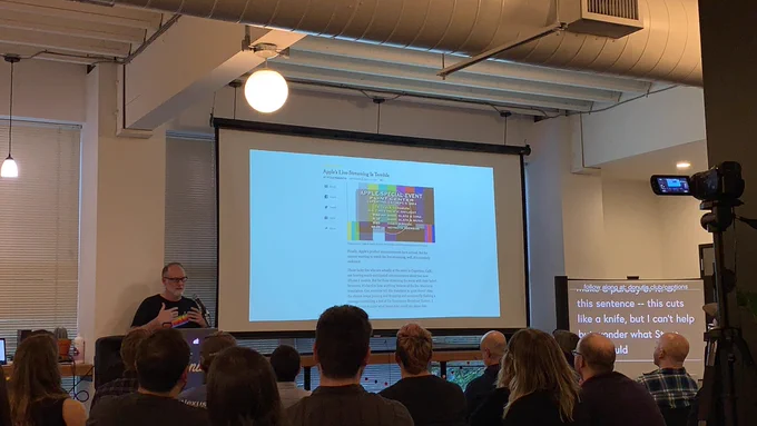 ‪A person giving a talk at a podium in front to a room full of people in chairs. Behind the speaker is a projector screen with an article titled “Apple’s Live Streaming Is Trouble.”‬