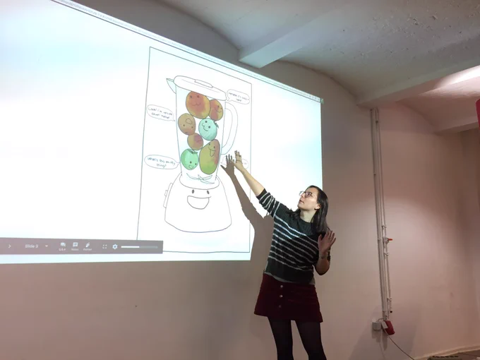 Yulia on stage and pointing at a slide. The slide has a blender on it to represent the mix of principles in JavaScript.