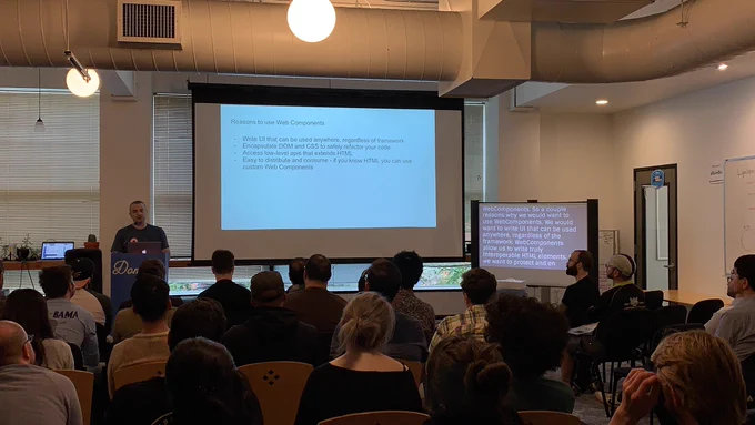 A person giving a talk at a podium in front to a room full of people in chairs. Behind the speaker is a projector screen with a list of reasons to use Web Components.