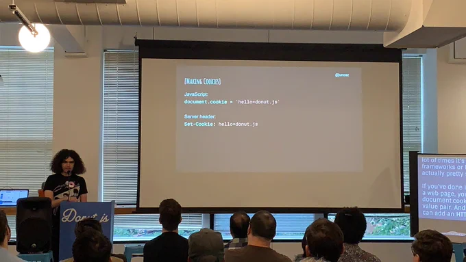 A person giving a talk at a podium in front to a room full of people in chairs. Behind the speaker is a projector screen with some JavaScript code on it.