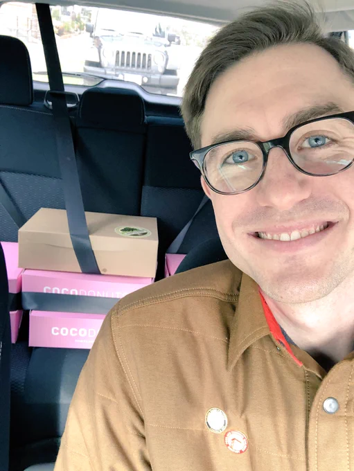 A selfie of me smiling in the driver’s seat with several boxes of donuts visible in the backseat.
