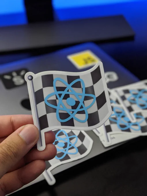 Me holding up a React Final Form (a checkered flag with React logo) sticker in front of my laptop which has other stickers on it.