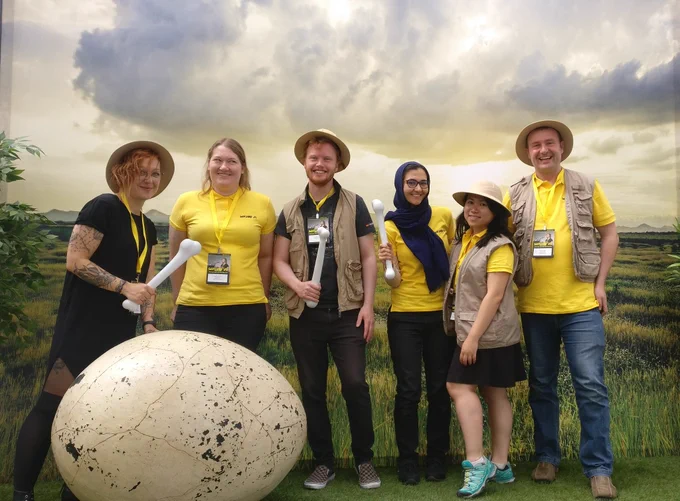 Five people dressed in explorer hats, with a huge Dino egg with a scenic background