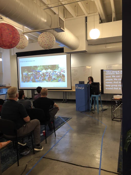 A woman giving a talk about urban planning in front of an audience. A picture is on the projection screen next to her of a crowd of people with the caption “Design for optimal group size”