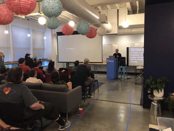 A person giving a talk next to a projection screen on which are the words “co = checkout.” About 20 people sitting in front watching the talk.