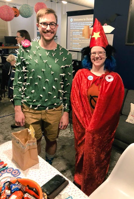 Two people in costumes for Halloween standing at the check-in table. The first person has a green shirt with white pipe cleaners and a paper flower on it. The second person has a shirt with a zero on it, a wizard’s cape and hat with a sword through it, and cat ears.