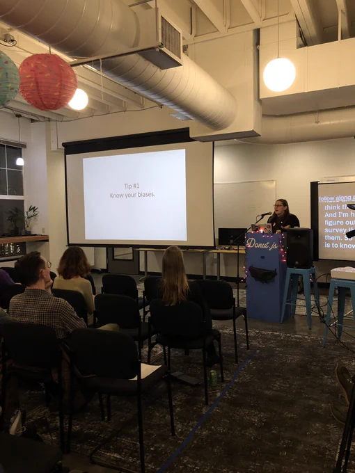 A person giving a presentation to a room full of people. There is a slide on a screen that reads “Tip #1: know your biases”