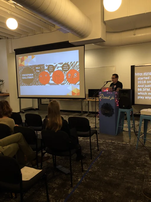 A person giving a presentation at a podium to an audience in chairs. On a projection screen is the logo of the community radio station he works for.