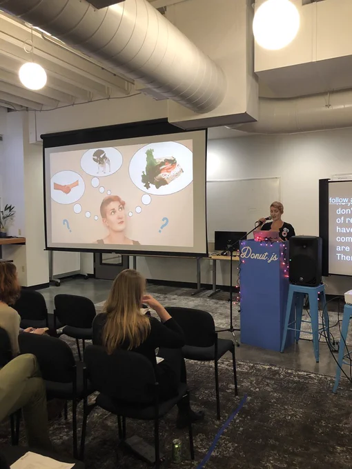 A person giving a presentation at a podium in front of an audience. There is a slideshow screen showing a picture of that same person with three thought bubbles containing a pair of shaking hands, a dog dressed up as a dinosaur, and a sandwich.