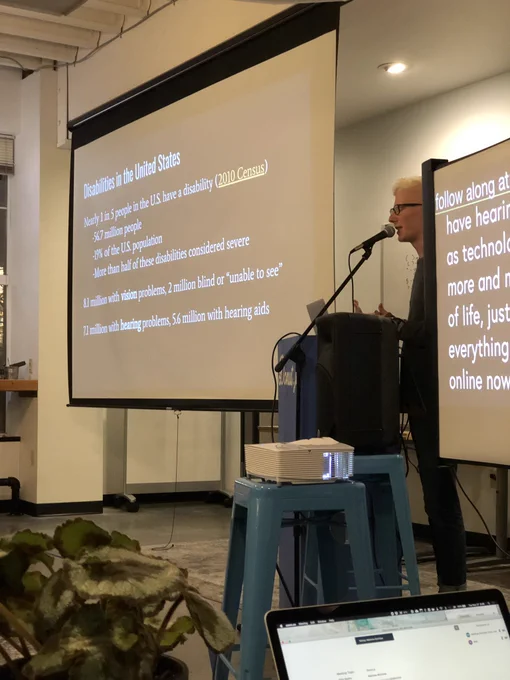 A person standing at a podium giving a talk. There is a projection screen with a slide outlining statistics about how many disabled people are in the US.