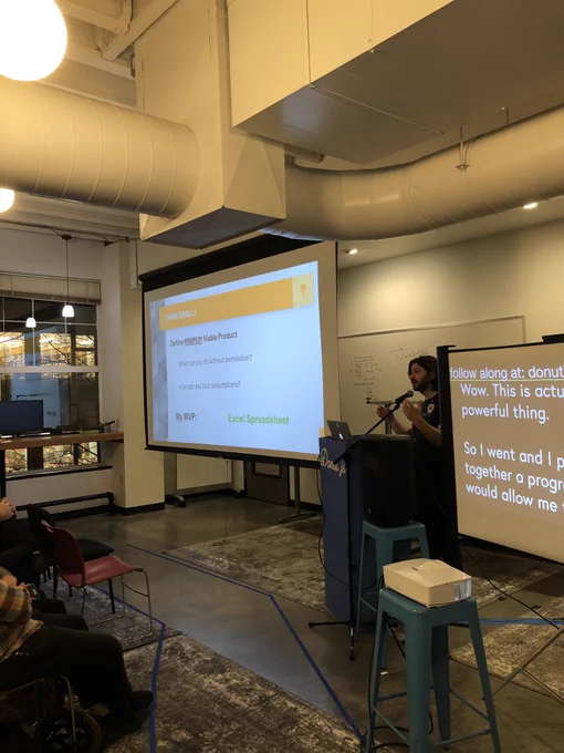 A person standing at a podium giving a talk. There is a projection screen with a slide explaining the importance of a minimum viable product and giving an Excel spreadsheet as an example.