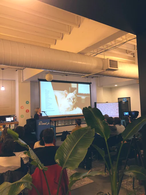 A woman is standing at a podium giving a talk in front of a room full of people sitting. A projection screen to her right has a large photo of a cat and a list of distractions.