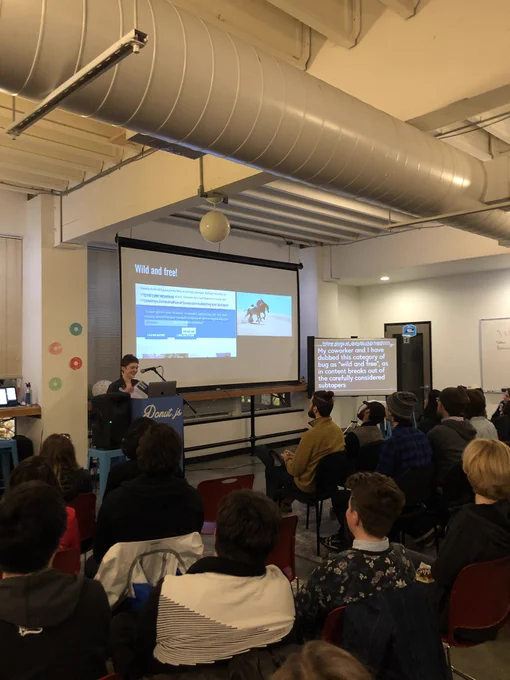 A woman giving a talk at a podium in front of a room of people. There is a projector showing a messed up browser layout next to a photo of a wild horse.