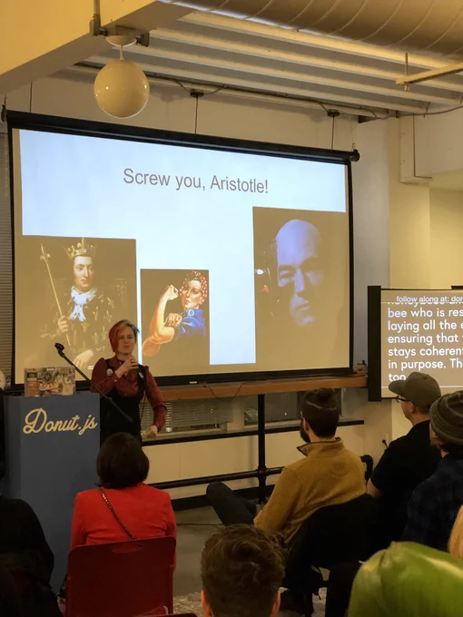 A woman giving a talk in front to a room full of people.  Behind her is a projector screen with a picture of a king (representing the mistaken early conception of the queen bee as a ‘king’), the ‘We Can Do It’ picture (representing the worker bee), and Jean Luc-Picard as a borg (representing the drone bee).