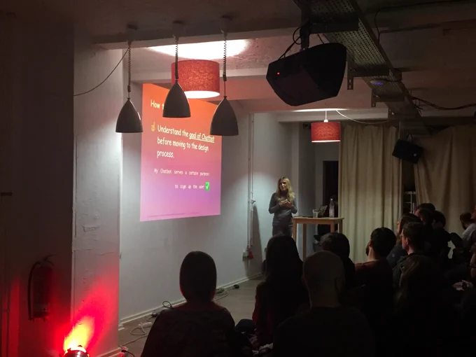 Ilona standing in front of audience. She’s moving her hands and there is a red slide with text behind her.