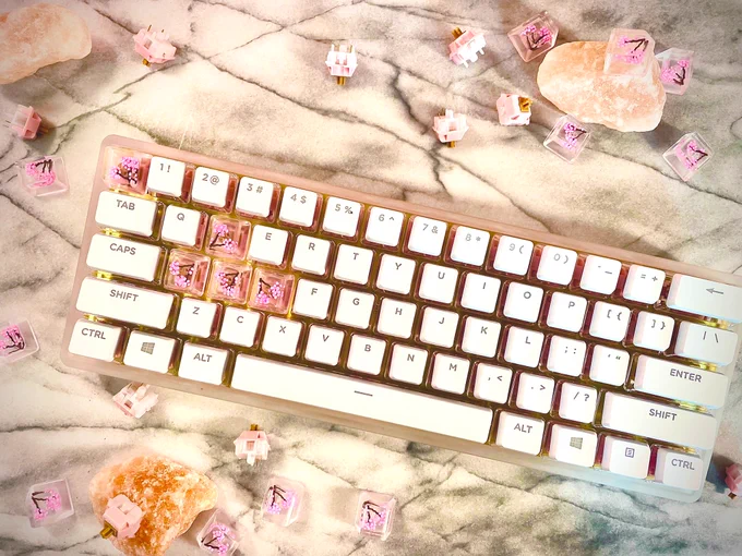 A keyboard sits on a white marble platter with rose quartz, keycaps, and key switches surrounding it. The case is frosted acrylic, and the escape and WASD keys are clear acrylic with hand-made cherry blossoms. The remaining keys are white with a crystal base.