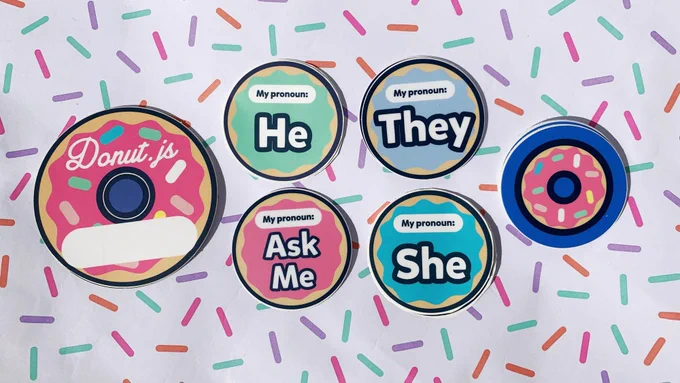 Donut-shaped name tag stickers, Donut.js logo stickers, and several pronoun stickers (Ask Me, He, She, They) arranged on paper patterned with sprinkles.
