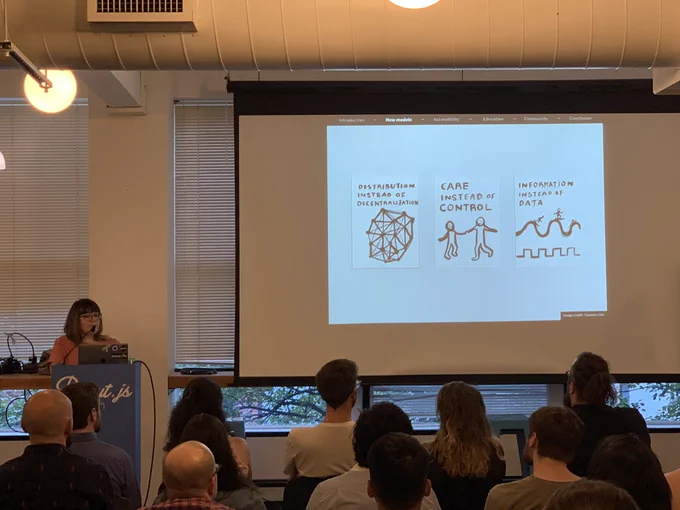 A person giving a talk at a podium in front to a room full of people in chairs. Behind the speaker is a projector screen with three illustrations focusing on internet communities.