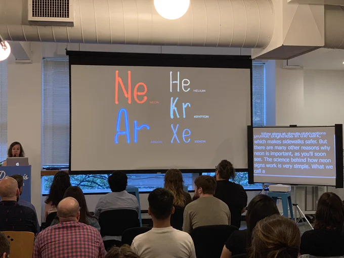 A person giving a talk at a podium in front to a room full of people in chairs. Behind the speaker is a projector screen listing the five noble gasses that are used in neon lights.