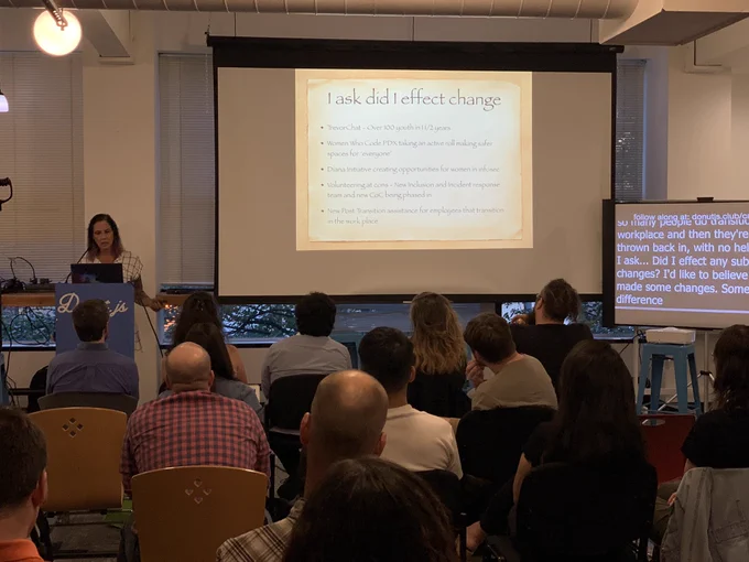 A person giving a talk at a podium in front to a room full of people in chairs. Behind the speaker is a projector screen with a slide that reads “I ask: did I effect change?” with a list of the speakers’ volunteer accomplishments over the last year.