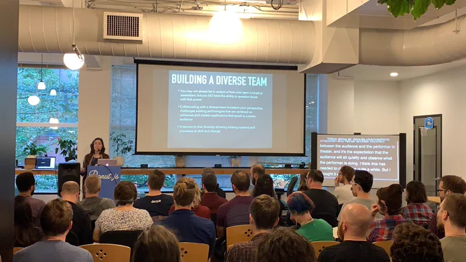 A person giving a talk at a podium in front to a room full of people in chairs. Behind the speaker is a projector screen with ‘Building a Diverse Team’ written on it.
