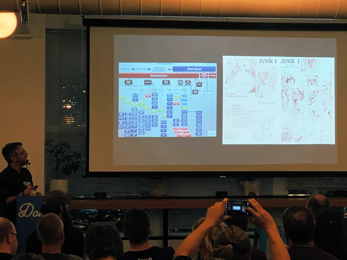 ‪A person giving a talk at a podium in front to a room full of people in chairs. Behind the speaker is a projector screen with ‬a diagram of a banking system phone tree next to a diagram of a text adventure. The comparison shows their similarity.