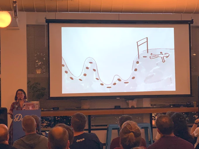 ‪A person giving a talk at a podium in front to a room full of people in chairs. Behind the speaker is a projector screen with ‬an illustration of a person lying down after running a race.