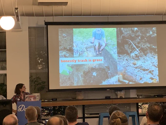 ‪A person giving a talk at a podium in front to a room full of people in chairs. Behind the speaker is a projector screen with ‬a photo of her digging up a trash pit with the caption “honestly trash is gross.”