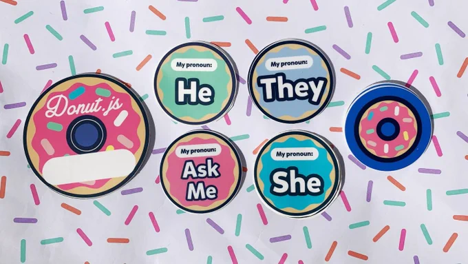 Donut-shaped name tag stickers, Donut.js logo stickers, and several pronoun stickers (Ask Me, He, She, They) arranged on paper patterned with sprinkles.