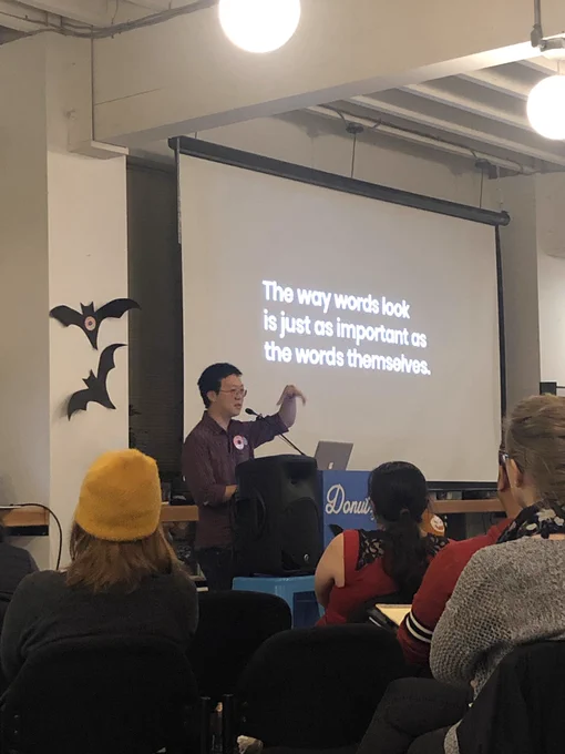 A person giving a talk at a podium in front to a room full of people in chairs. Behind the speaker is a projector screen with these words: “The way words look is just as important as the words themselves.”