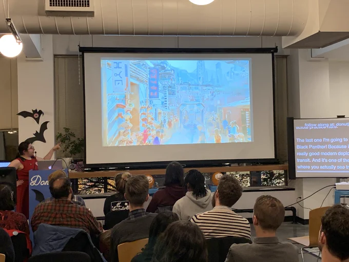 A person giving a talk at a podium in front to a room full of people in chairs. Behind the speaker is a projector screen with a film still from ‘Black Panther’ showing public transit in Wakanda.