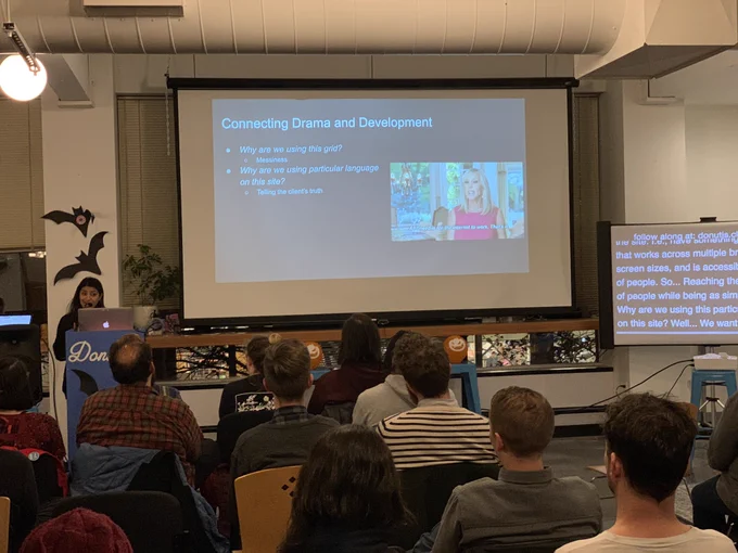 A person giving a talk at a podium in front to a room full of people in chairs. Behind the speaker is a projector screen with the title ‘Connecting Drama and Development.’