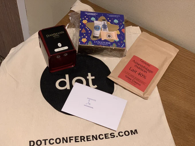 A dot conferences tote bag with the following items placed on it - a letter addressed to Ire, a pack of chocolate, a container of green tea, a pack with L'Occtitane products