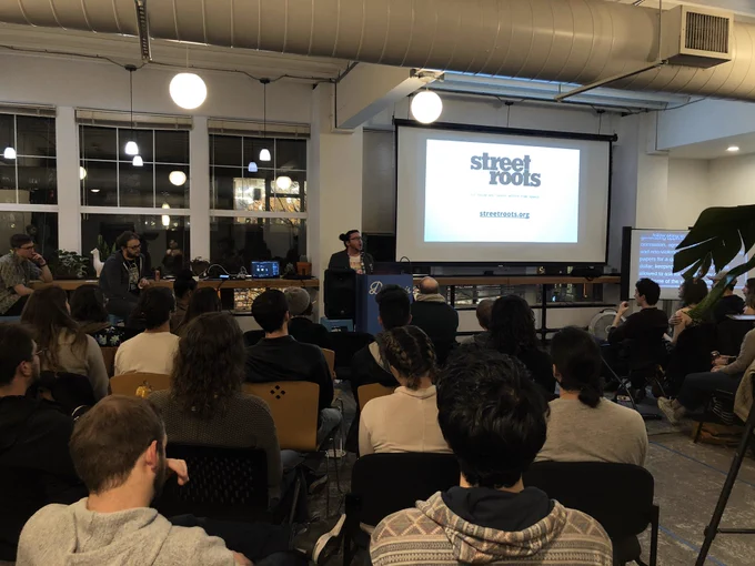 A person giving a talk at a podium in front to a room full of people in chairs. Behind the speaker is a projector screen with ‘Street Roots’ on it.