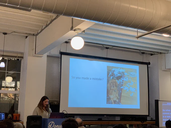 A person giving a talk at a podium in front to a room full of people in chairs. Behind the speaker is a projector screen with the words “so you made a mistake?” With a photo of the speaker’s dog standing front of a pillow ripped to shreds.