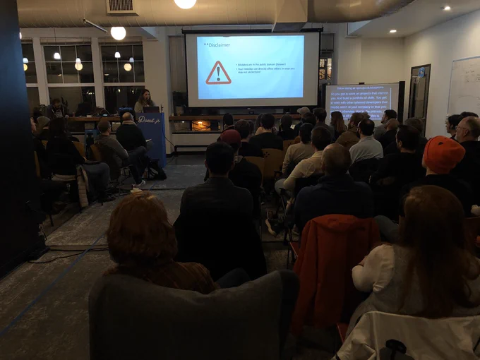 A person giving a talk at a podium in front to a room full of people in chairs. Behind the speaker is a projector screen with a warning sign on it and the words “mistakes in git can be in public forever. And they may affect others in unforeseen ways”