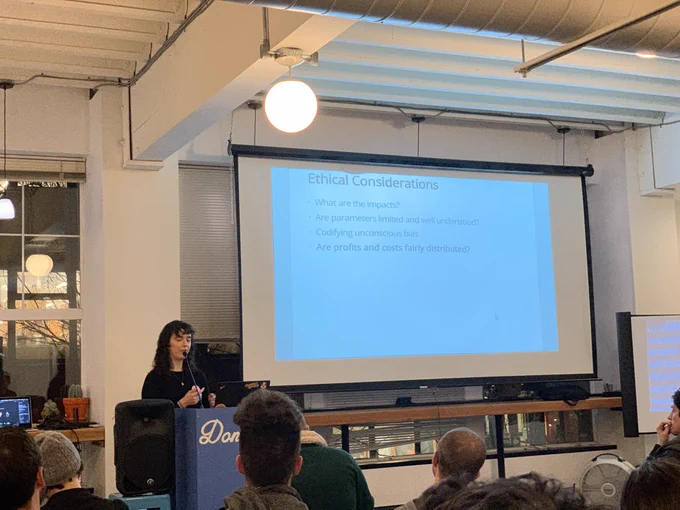 A person giving a talk at a podium in front to a room full of people in chairs. Behind the speaker is a projector screen with the words: 
ethical considerations:
- what are the impacts?
- are parameters limited and well understood?
- codifying unconscious bias
- **are profits and costs fairly distributed?**